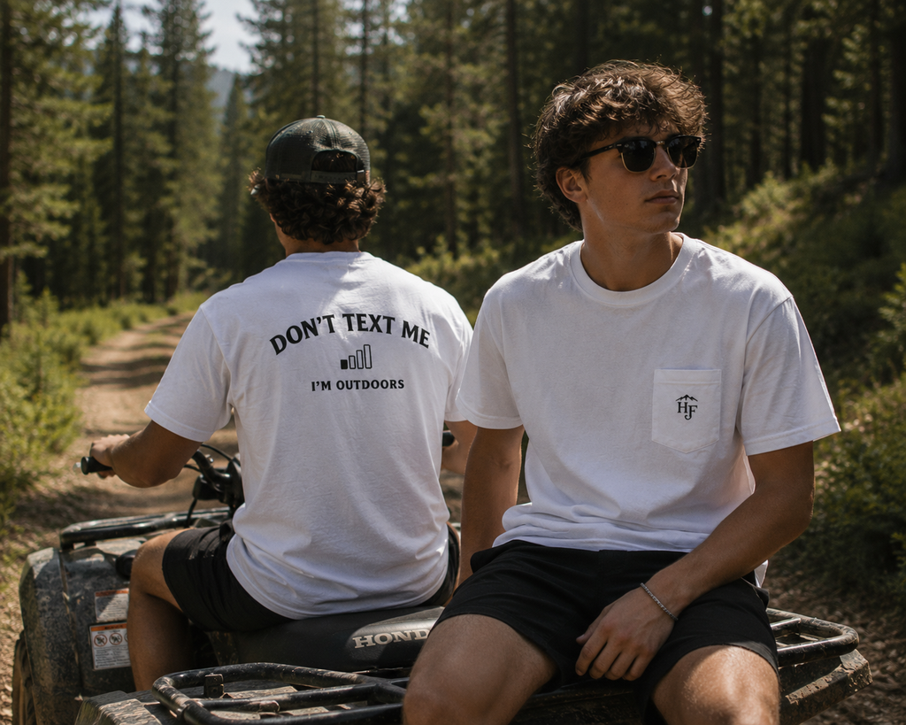 Two people wearing white "Don't Text Me I'm Outdoors" outdoor pocket tees on an ATV in a forest, designed for fishing, camping and outdoor adventure.