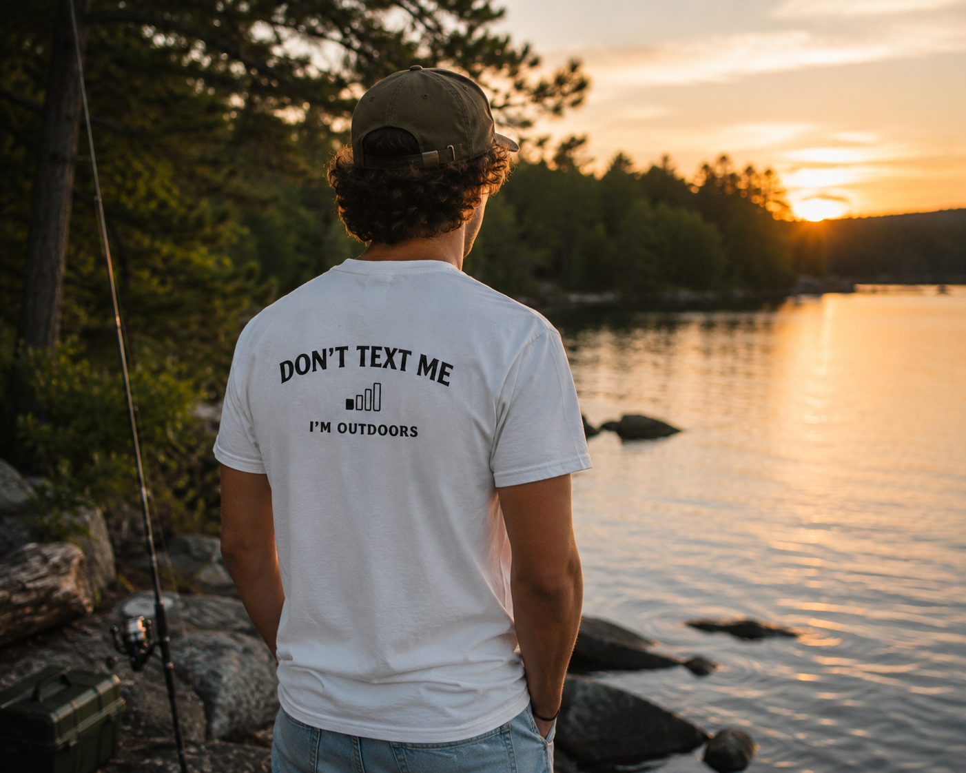 Person wearing white "Don't Text Me I'm Outdoors" outdoor pocket tee by a lake at sunset, designed for fishing and outdoor lifestyle.